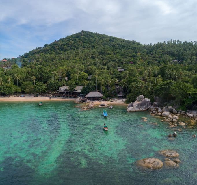 An aerial view of a tropical cove with clear turquoise water, small boats, a sandy beach, and a lush green, tree-covered hill.