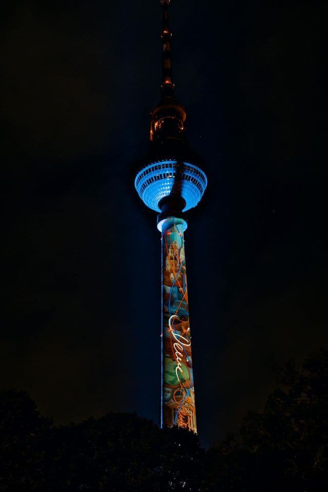 Una torre alta dalla cima sferica è illuminata da luce blu e proiezioni colorate contro il cielo notturno scuro.