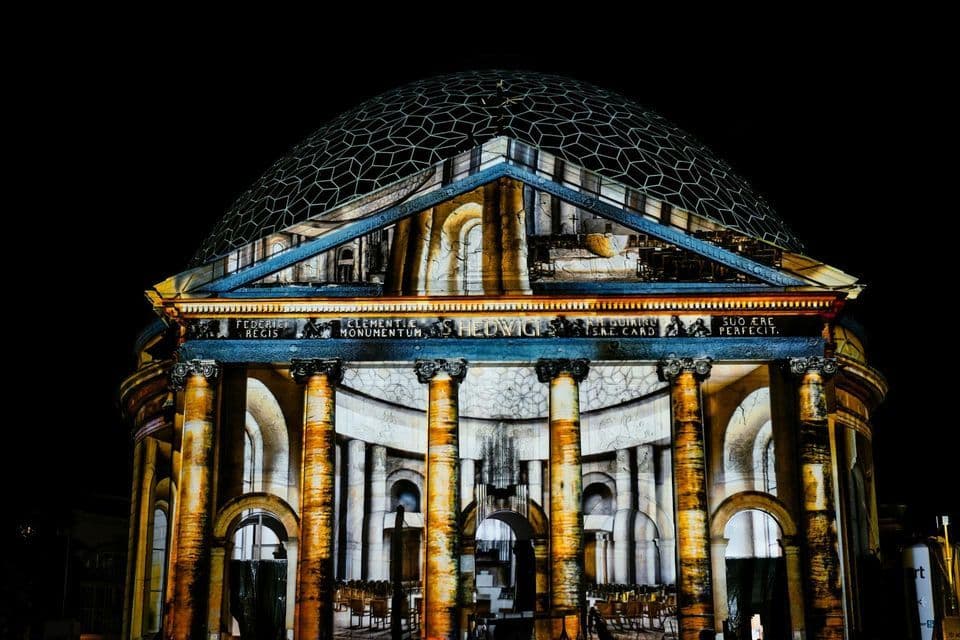 Un grande edificio classico con cupola e colonne, illuminato di notte da una proiezione luminosa che ne rivela la struttura interna.
