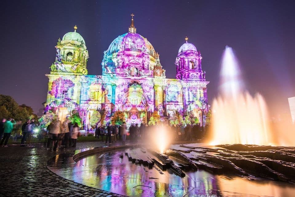 Di notte, un edificio a cupola riccamente ornato è illuminato da luci colorate proiettate, con una fontana e una folla di persone in primo piano.