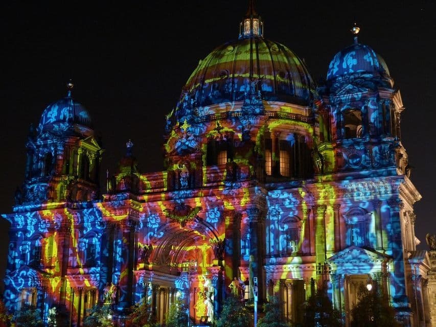 Un edificio a cupola riccamente decorato, illuminato da proiezioni di luce colorate blu, gialla e rossa, su uno sfondo di cielo notturno scuro.