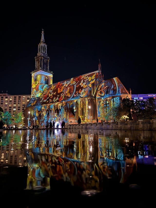 Una chiesa è illuminata di notte da una proiezione di farfalle colorate, con il suo riflesso visibile sull'acqua in primo piano.
