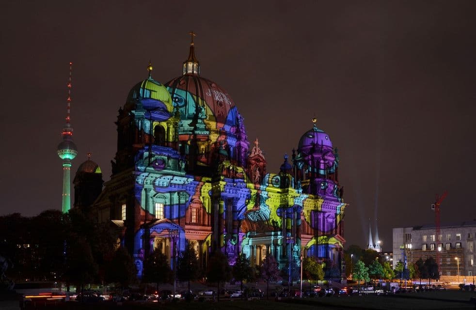 Un grande edificio a cupola è illuminato di notte da proiezioni di luce colorate, con un'alta torre televisiva illuminata in lontananza.