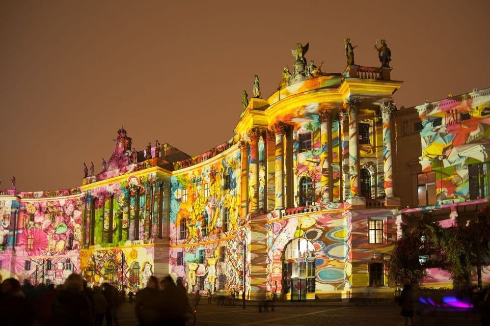 Una facciata di edificio riccamente decorata, illuminata di notte con un vivace e colorato spettacolo di luci proiettate, con la gente che osserva dal basso.