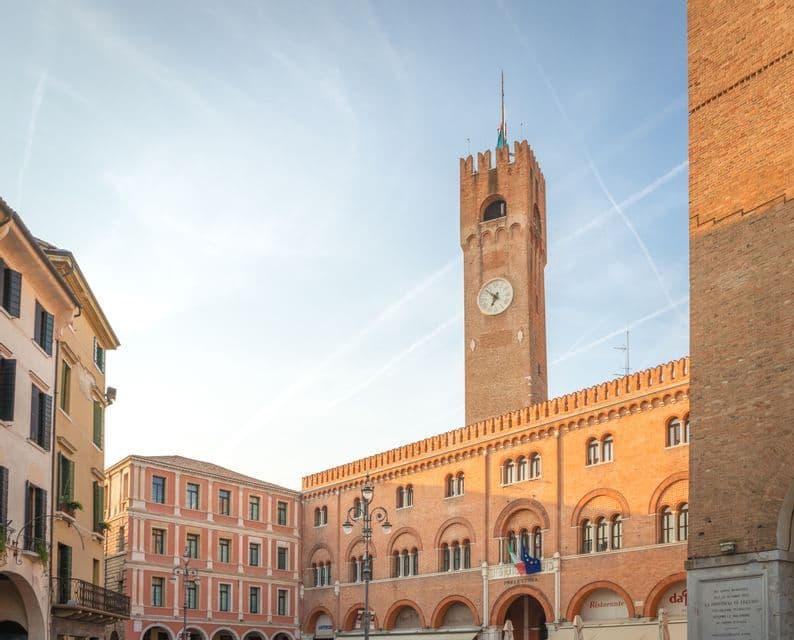 Ein sonnenbeschienener, roter Backsteinglockenturm und ein angrenzendes Bogengebäude stehen auf einem historischen Marktplatz unter einem hellblauen Himmel.
