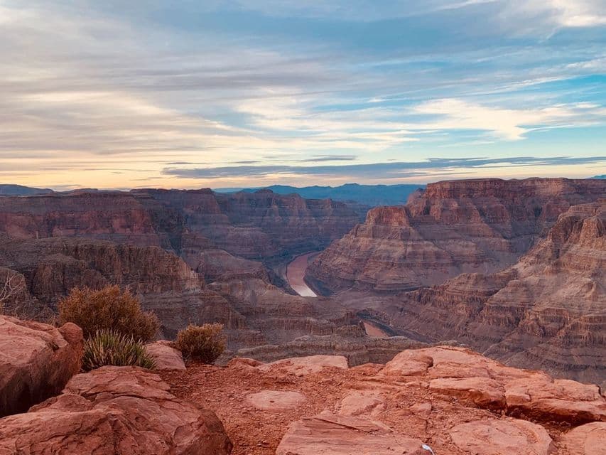 Ein weiter Blick auf einen tiefen Canyon mit einem sich schlängelnden Fluss, von einem felsigen Aussichtspunkt unter einem bewölkten Himmel bei Sonnenuntergang.