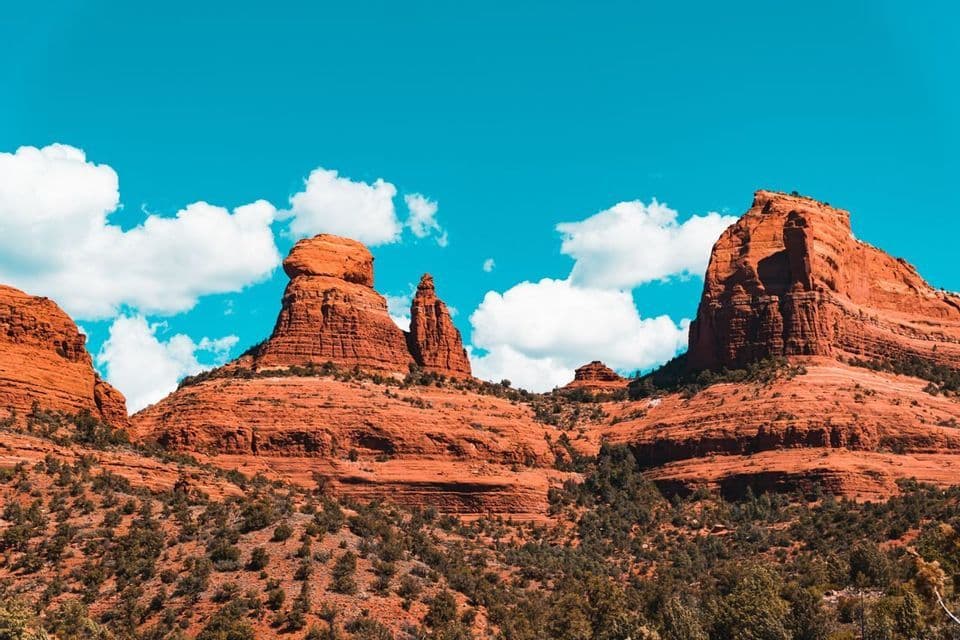 Large red rock formations rise from a landscape of green scrub under a bright blue sky with white clouds.