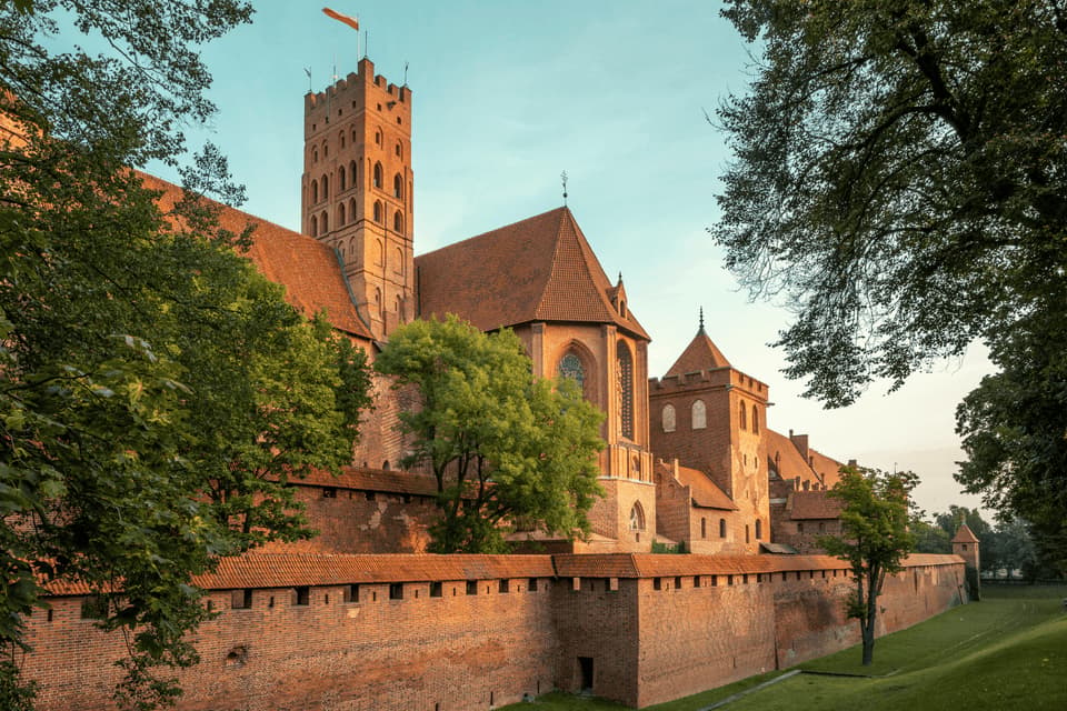 Un grande castello medievale in mattoni rossi con un'alta torre si erge dietro un muro fortificato, incorniciato da alberi verdi al tramonto.
