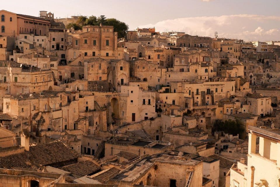 Antichi edifici in pietra color sabbia sono fittamente raggruppati su un fianco di collina, illuminati dalla calda luce del sole sotto un cielo velato.