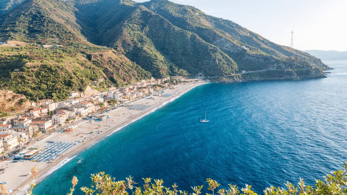 Veduta aerea di una località costiera con spiaggia sabbiosa ai piedi di montagne verdi, che si affaccia su un mare blu profondo con una barca a vela.
