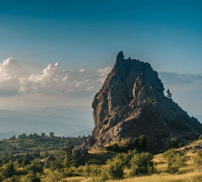 Una grande formazione rocciosa frastagliata si erge su una collina verde, con montagne lontane e velate sotto un cielo azzurro con qualche nuvola.