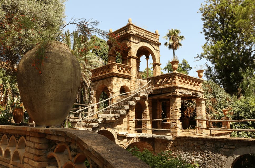 Un grande vaso di terracotta poggia su un muro di mattoni, affacciandosi su un'elaborata follia in mattoni a più livelli con archi, in un giardino lussureggiante.