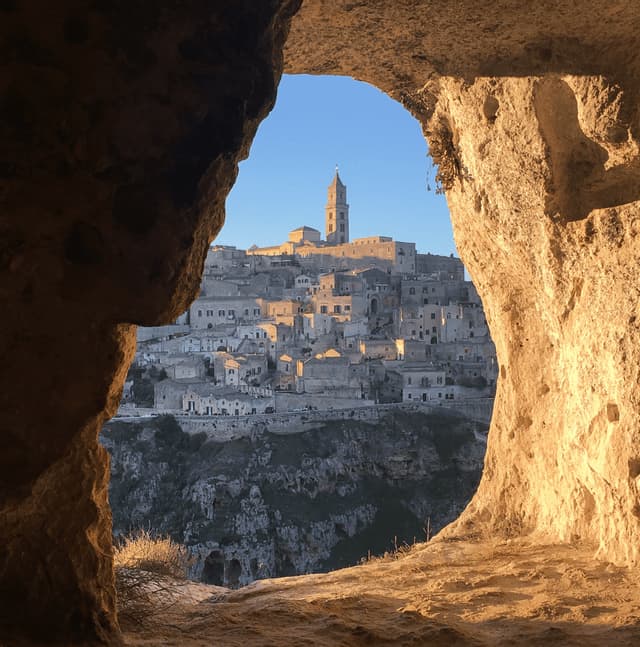 Una veduta di una storica città in pietra su un fianco di collina, ripresa dall'interno di un'apertura di grotta e illuminata dalla luce dell'ora d'oro.