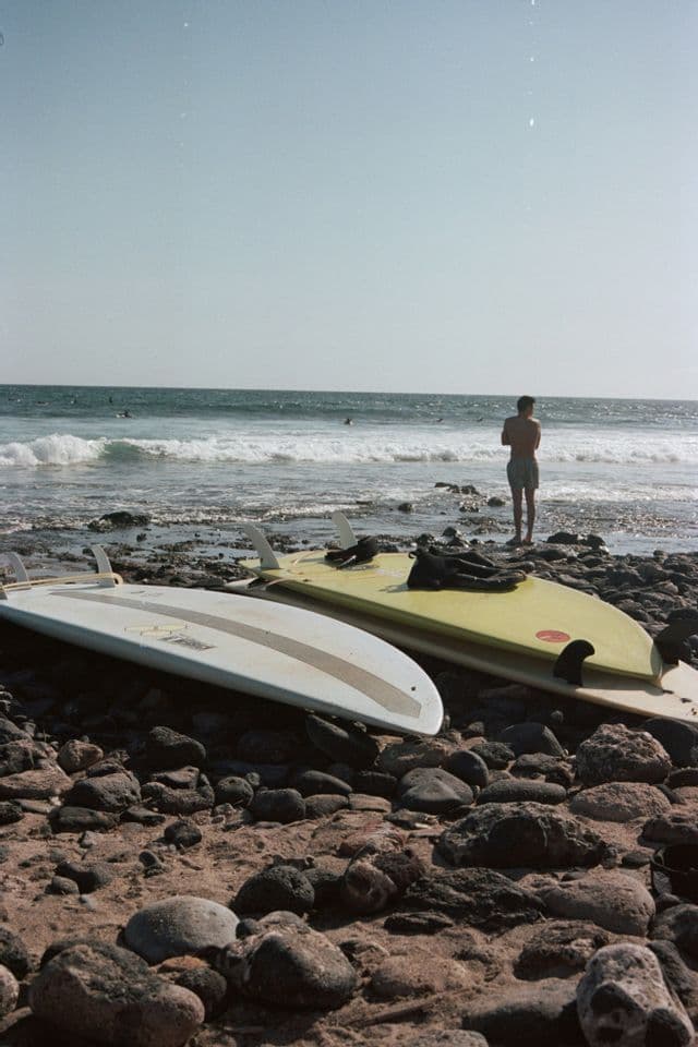 Due tavole da surf poggiano su una spiaggia rocciosa con una persona in piedi sul bagnasciuga che guarda l'oceano.