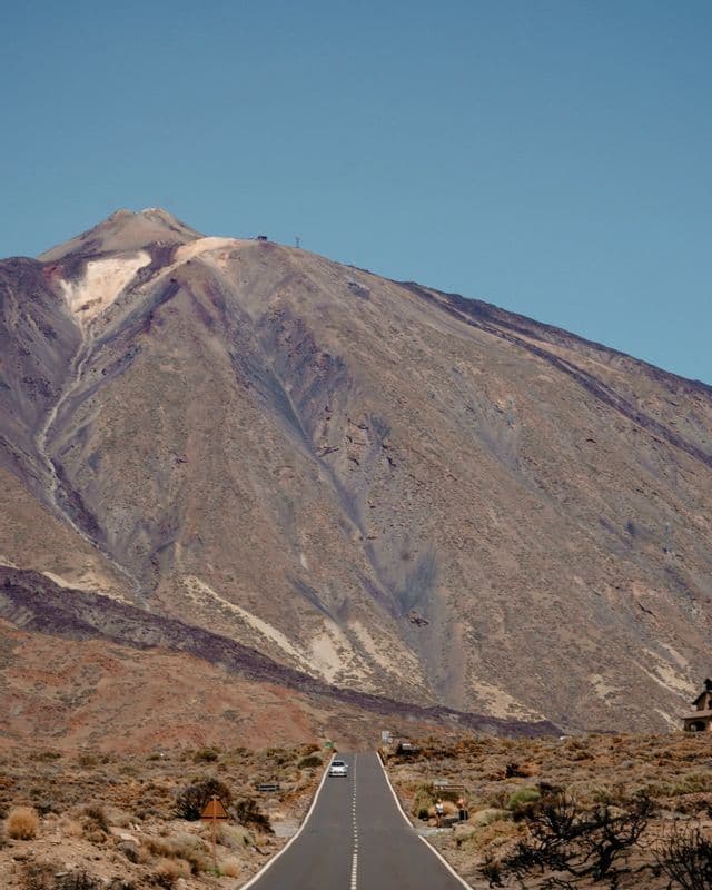 Un'auto bianca percorre una strada lunga e dritta attraverso un paesaggio arido, dirigendosi verso la base di un'imponente montagna vulcanica sotto un cielo blu limpido.