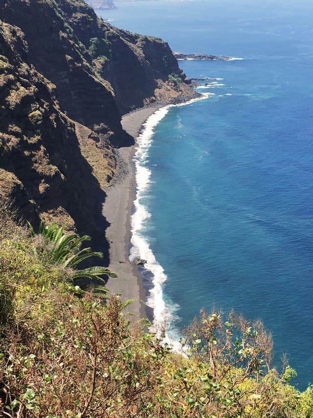 Vista aerea di una costa rocciosa, con una spiaggia di sabbia nera alla base di una scogliera e onde azzurre che si infrangono sulla battigia.