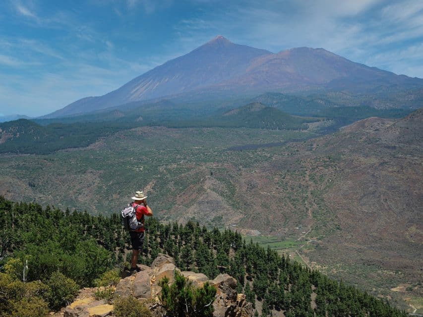 Un escursionista con cappello e zaino si trova su un punto panoramico roccioso, ammirando una valle boscosa verso un grande vulcano.