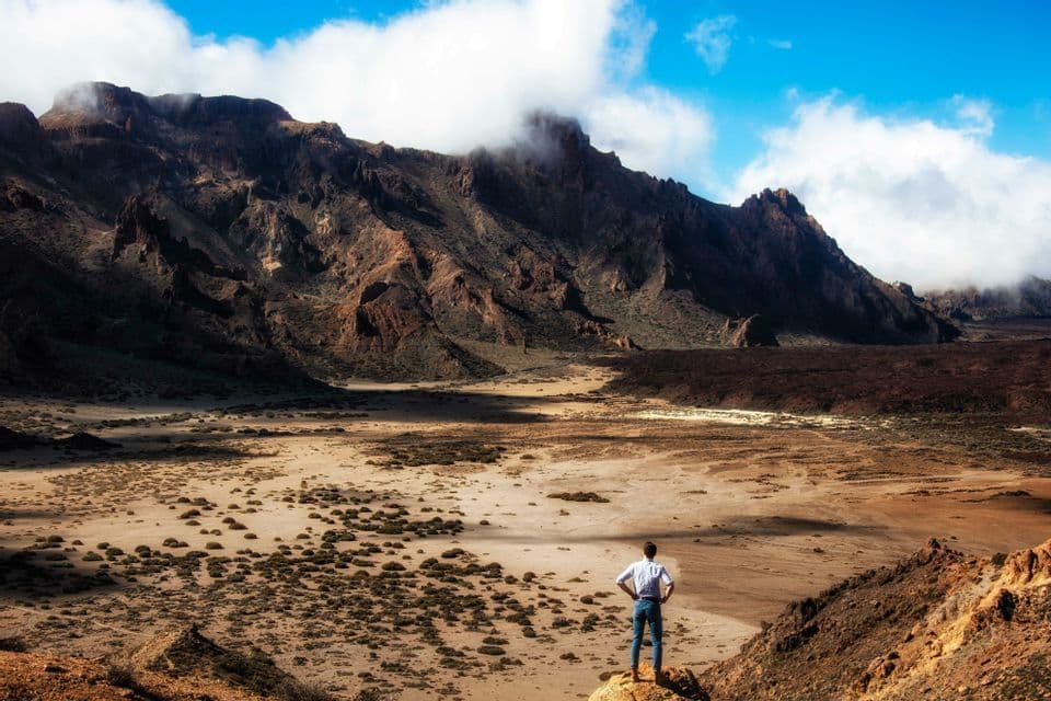 Un uomo in camicia bianca sta su uno sperone roccioso che domina una vasta valle arida con montagne aspre sullo sfondo.