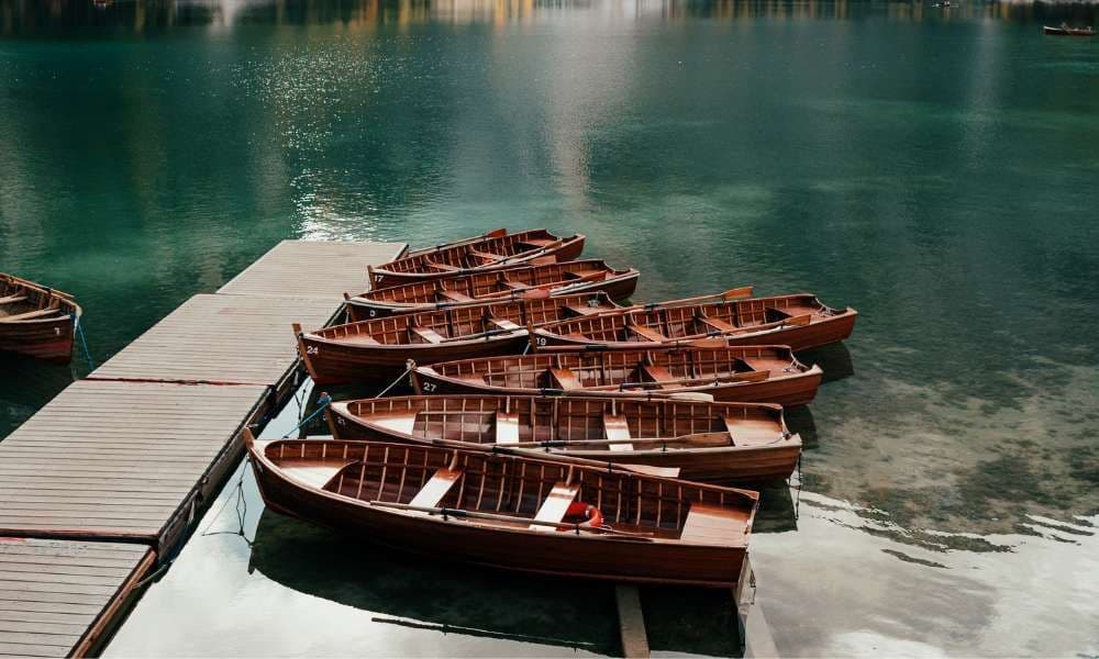 Eine Reihe leerer Holz-Ruderboote, vertäut an einem Holzsteg auf dem ruhigen, grünen Wasser eines Sees.