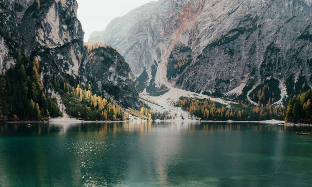 Ein ruhiger grüner Alpensee am Fuße steiler, felsiger Berge mit gelben und grünen Herbstbäumen.