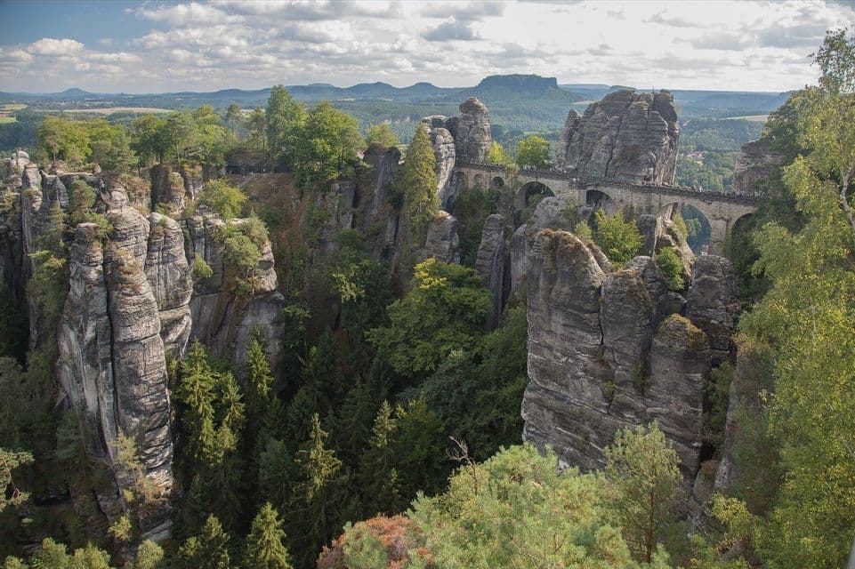 Eine gewölbte Steinbrücke verbindet hohe Felsformationen oberhalb eines grünen Tals, mit fernen Bergen unter einem teils bewölkten Himmel.