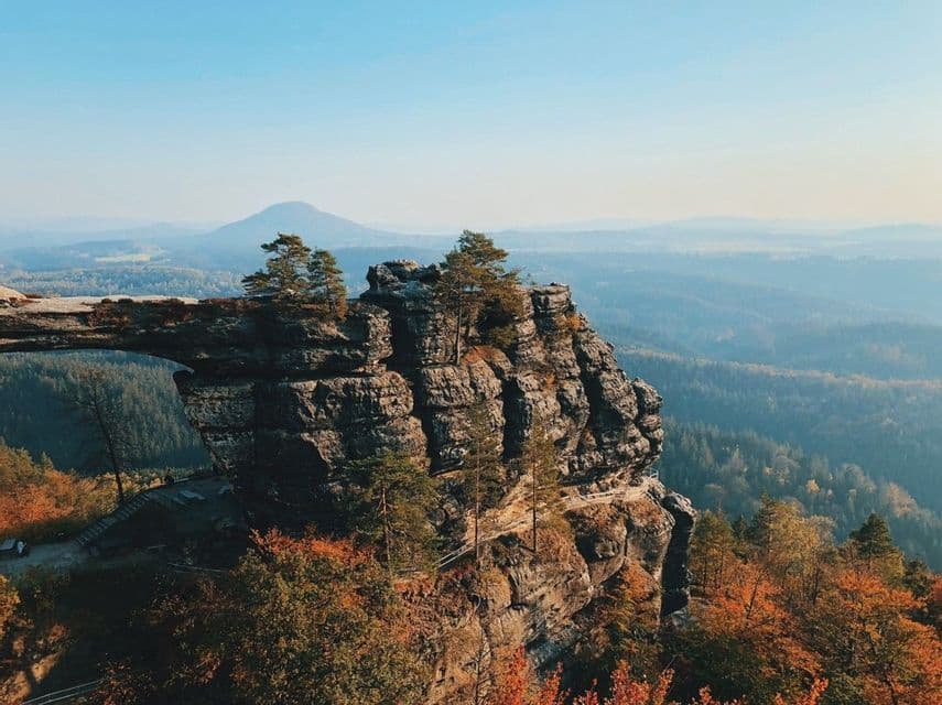 Ein natürlicher Steinbogen auf einer felsigen Klippe überblickt ein weites Tal mit Wäldern in Herbstfarben und fernen, dunstigen Bergen.