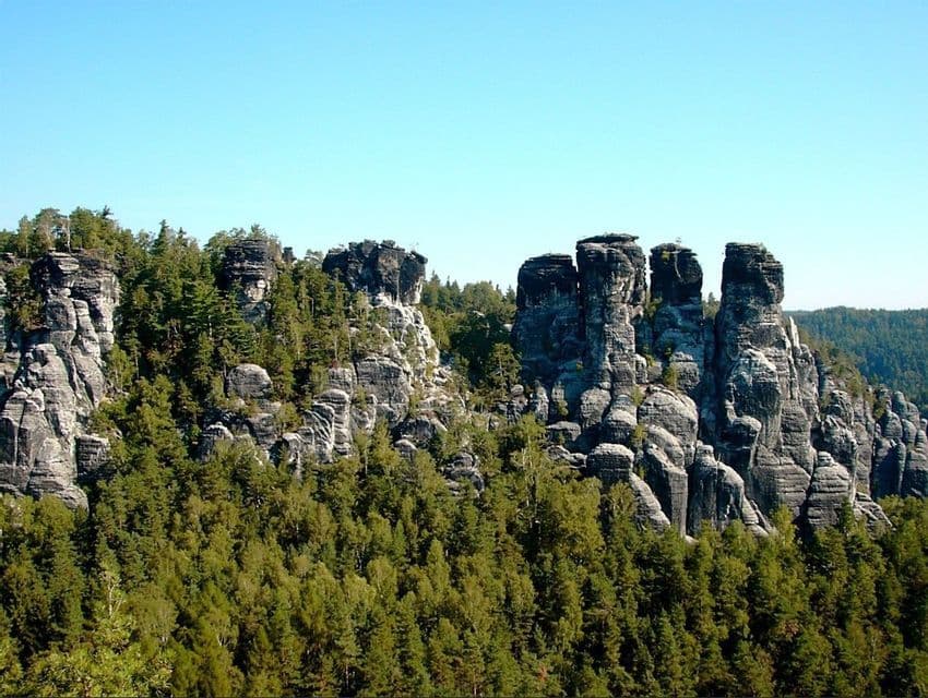 Hohe, von Bäumen bewachsene Felsformationen ragen aus einem dichten grünen Wald unter einem klaren blauen Himmel empor.