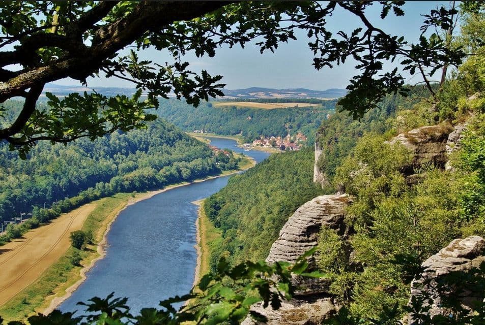 Blick von einem hohen Felsvorsprung auf einen breiten Fluss, der sich durch ein üppiges, bewaldetes Tal mit einem kleinen Dorf schlängelt.