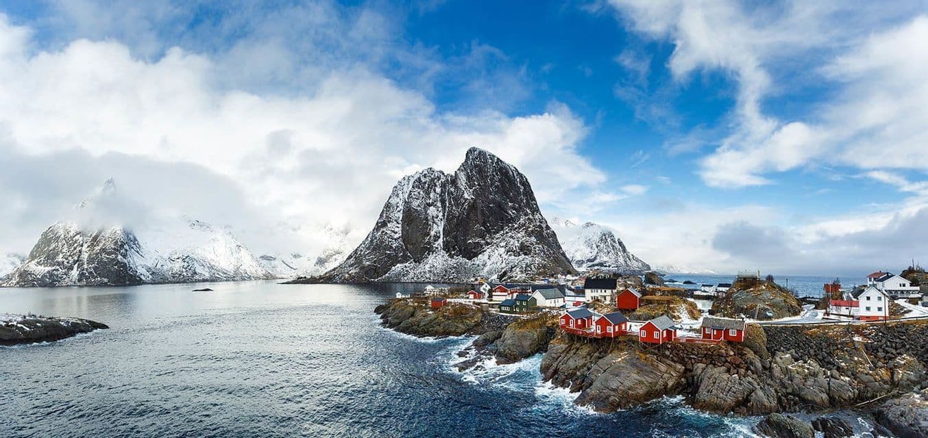A coastal village with red houses sits on a rocky shoreline by a fjord, with a large, snow-covered mountain behind it.