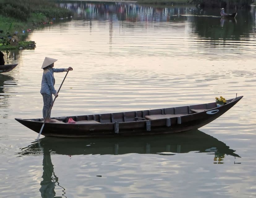 Una persona con sombrero cónico está de pie en un largo bote de madera, remando en aguas tranquilas y reflectantes con un solo remo.