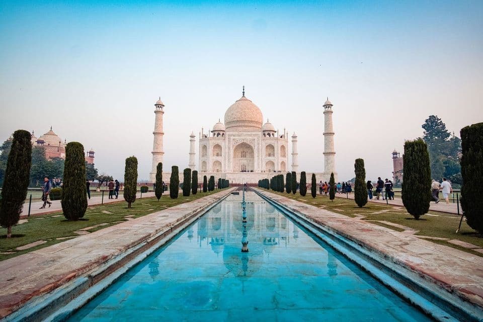 Il Taj Mahal si riflette in una lunga piscina fiancheggiata da giardini con cipressi, con visitatori che passeggiano sui sentieri.