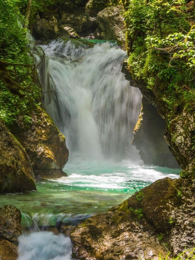 Ein Wasserfall ergießt sich in ein türkisfarbenes Becken, umrahmt von sonnenbeschienenen, moosbewachsenen Felsen und grünem Laub.