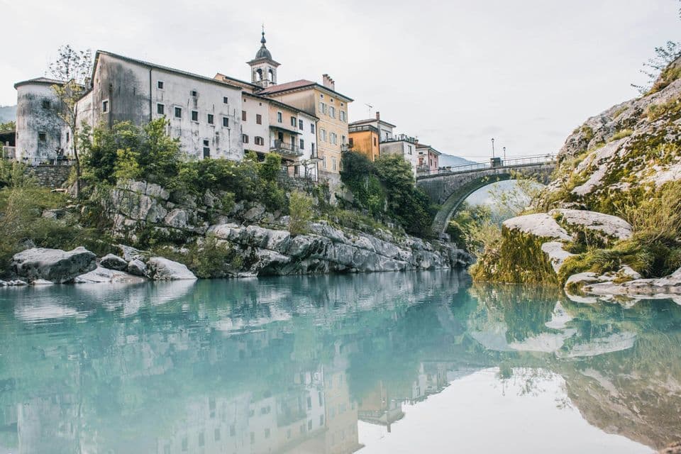 Alte Gebäude an einem felsigen Flussufer mit einer steinernen Bogenbrücke, deren Spiegelungen im ruhigen, türkisfarbenen Wasser sichtbar sind.