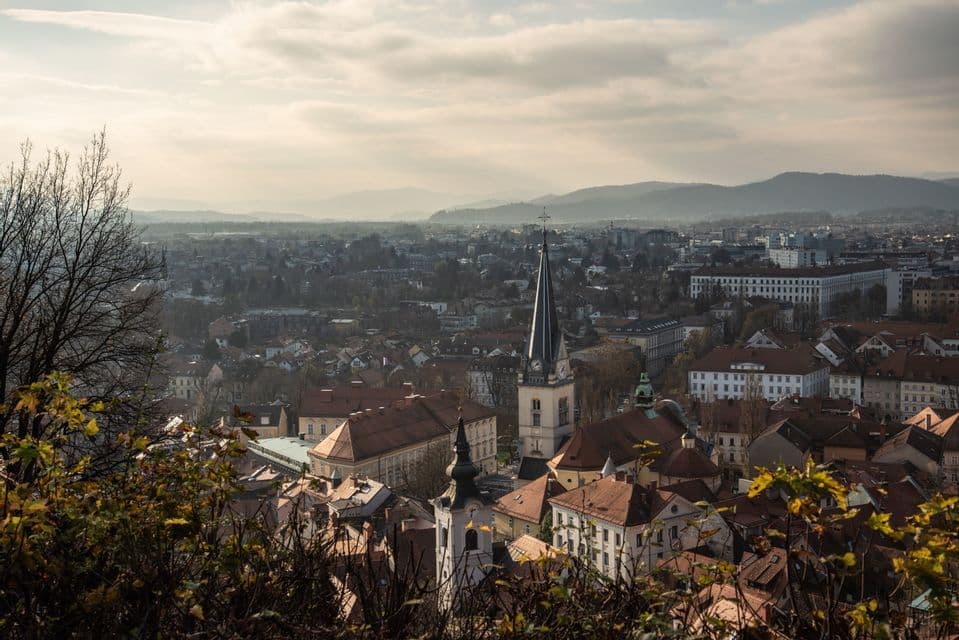Vogelperspektive einer Stadt mit roten Ziegeldächern und einem Kirchturm, diesige Berge in der Ferne unter bewölktem Himmel.