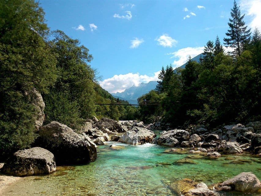 Zwei Personen radeln über eine Hängebrücke über einen klaren türkisfarbenen Fluss, der durch ein grünes, bergiges Tal fließt.