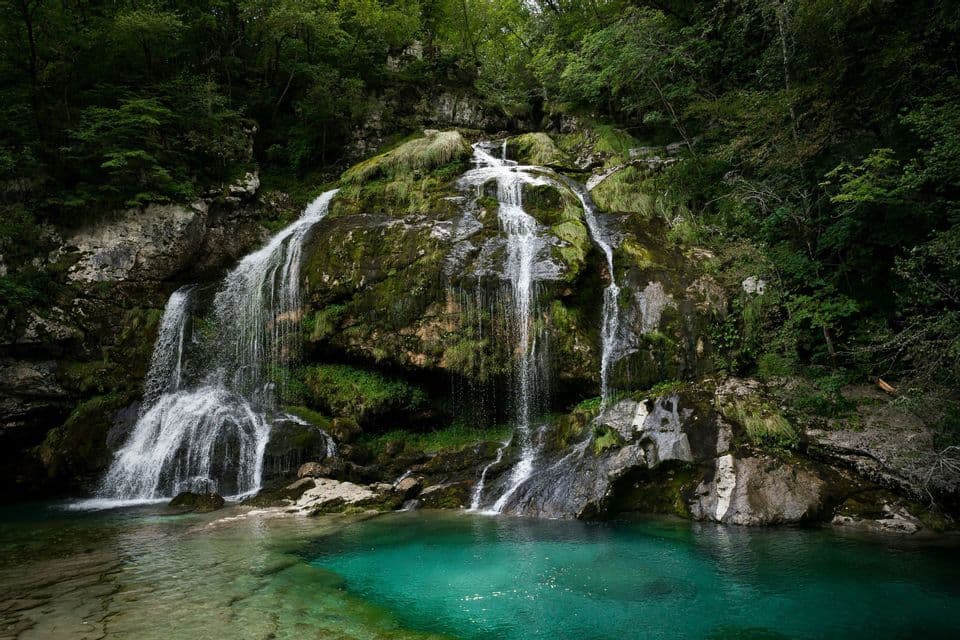 Ein breiter Wasserfall stürzt über moosbewachsene Felsen in ein klares türkises Becken am Fuße eines üppig grünen Waldes.