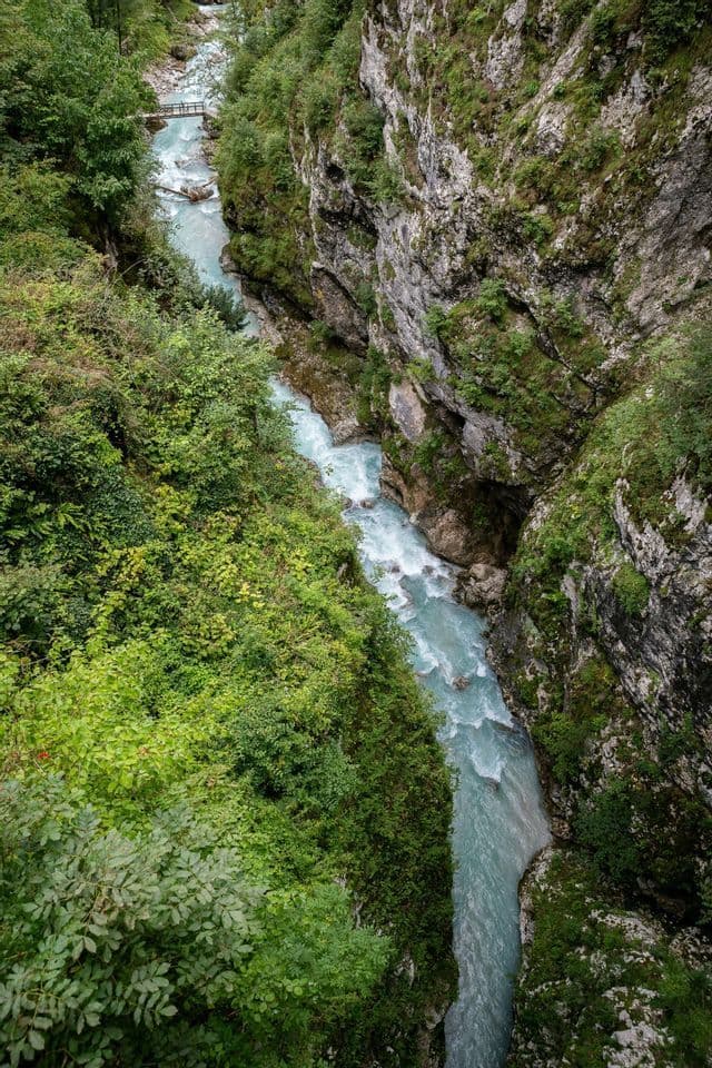 Eine Luftaufnahme eines türkisfarbenen Flusses, der sich durch eine tiefe, felsige Schlucht mit üppig grünen Bäumen und einer entfernten Holzbrücke schlängelt.