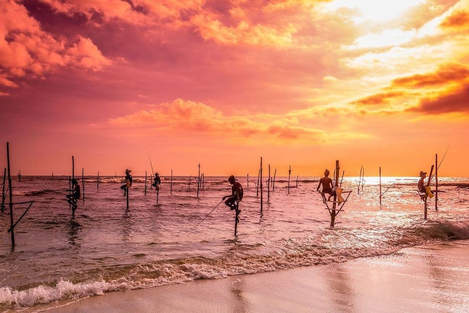 Mehrere Fischer sitzen auf hohen Holzstelzen im flachen Meer und halten Angelruten unter einem dramatischen, farbenfrohen Sonnenuntergangshimmel.