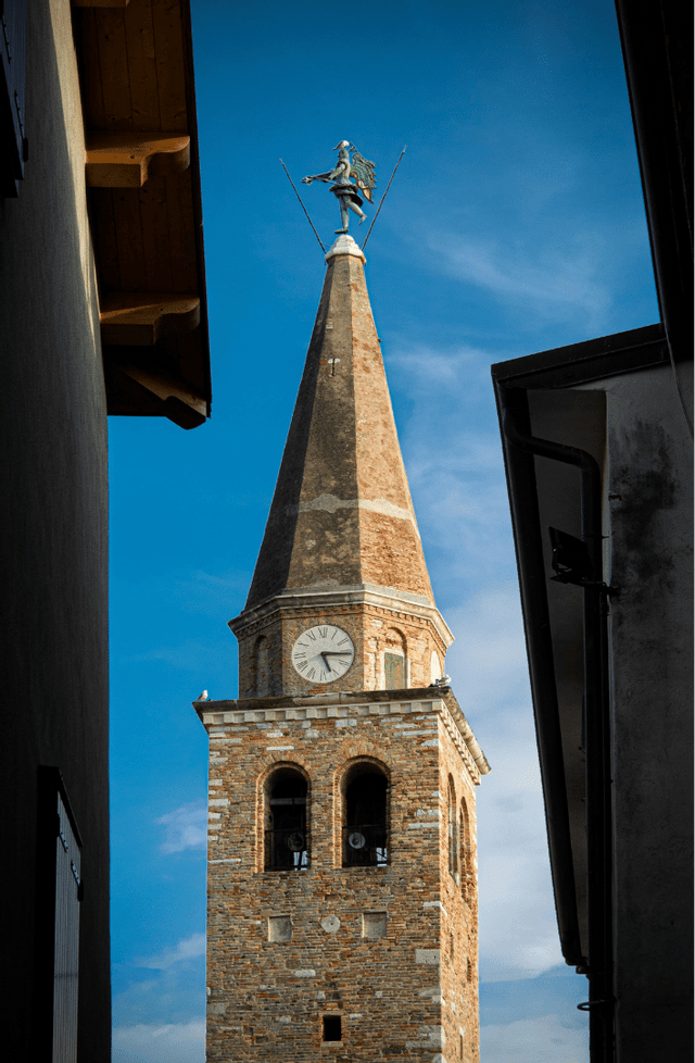 Una torre dell'orologio in pietra con una guglia e una banderuola a forma di angelo, incorniciata tra due edifici su uno sfondo di cielo blu.