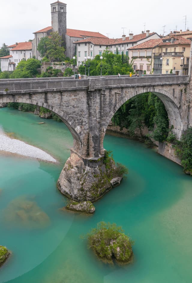 Un ponte ad arco in pietra attraversa un fiume turchese, con una città storica e una torre di pietra visibili sulla riva sullo sfondo.