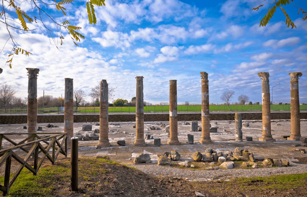 Una fila di antiche colonne di pietra si erge in un sito archeologico sotto un cielo azzurro parzialmente nuvoloso.