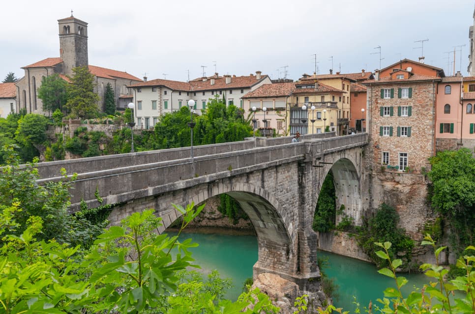 Un grande ponte ad arco in pietra attraversa un fiume turchese di fronte a una città collinare con edifici tradizionali e un campanile.