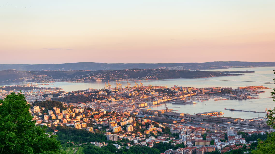 Una vista dall'alto di una città costiera con un grande porto, vista da una collina al tramonto sotto un cielo pastello.