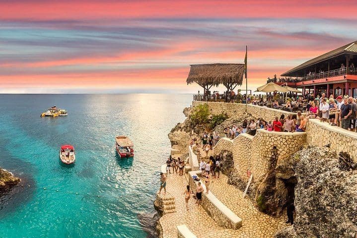 Una multitud de personas se reúne en un restaurante en el acantilado y en terrazas de piedra para ver la puesta de sol sobre un mar turquesa con barcos.