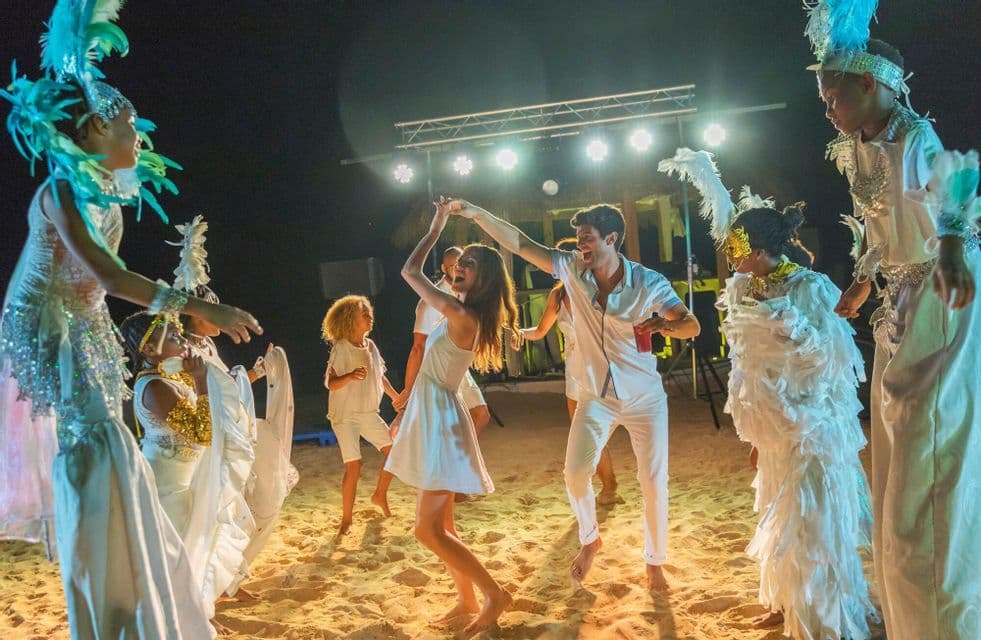 Un viaje en grupo de WeRoad bailando en una playa de arena por la noche con artistas luciendo elaborados trajes blancos y de plumas.