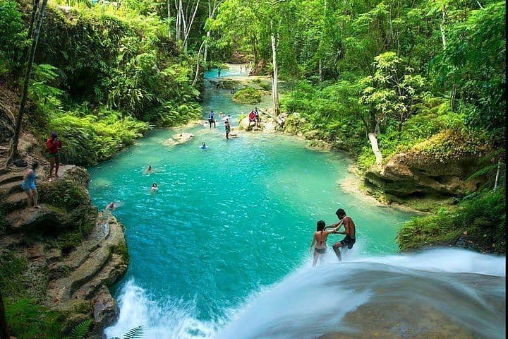 Un viaje en grupo de WeRoad nada en un río turquesa, con una pareja al borde de una cascada, rodeados de una selva exuberante.