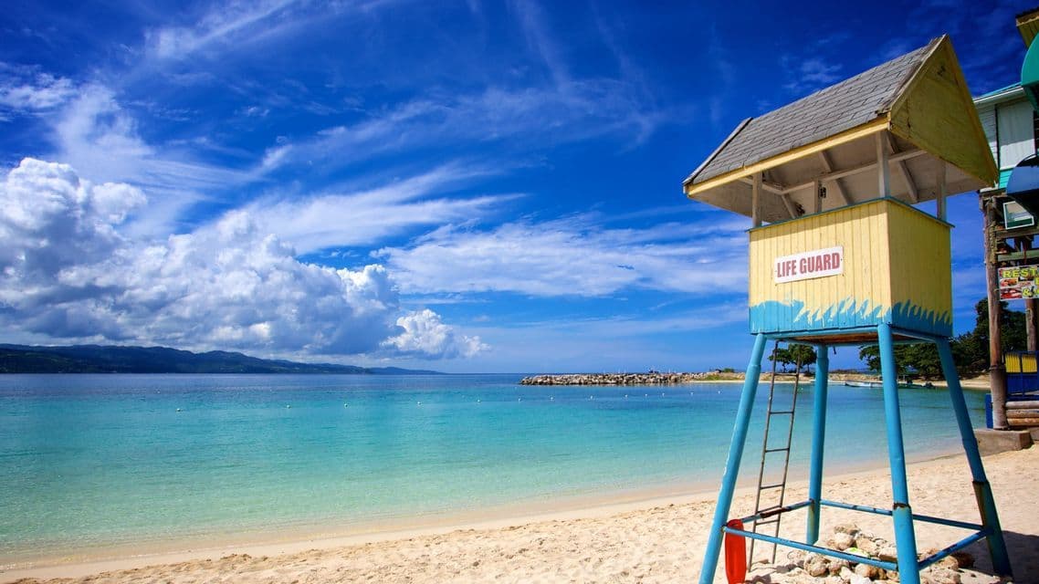 Una torre de salvavidas amarilla y azul se alza en una playa de arena con vistas a aguas turquesas bajo un cielo azul con nubes blancas.