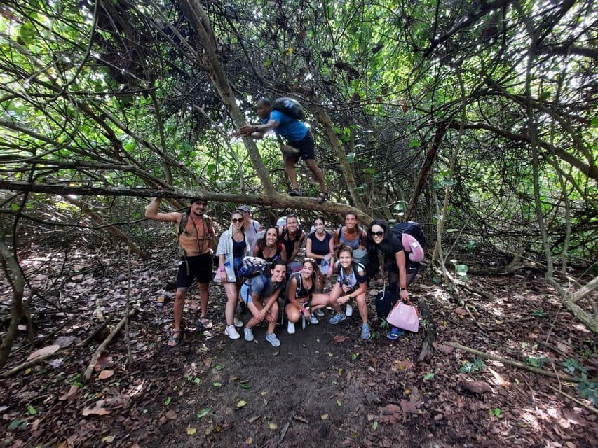Un groupe WeRoad en voyage avec sacs à dos pose pour une photo sur un sentier sous des branches d'arbres enchevêtrées, au cœur d'une forêt dense.