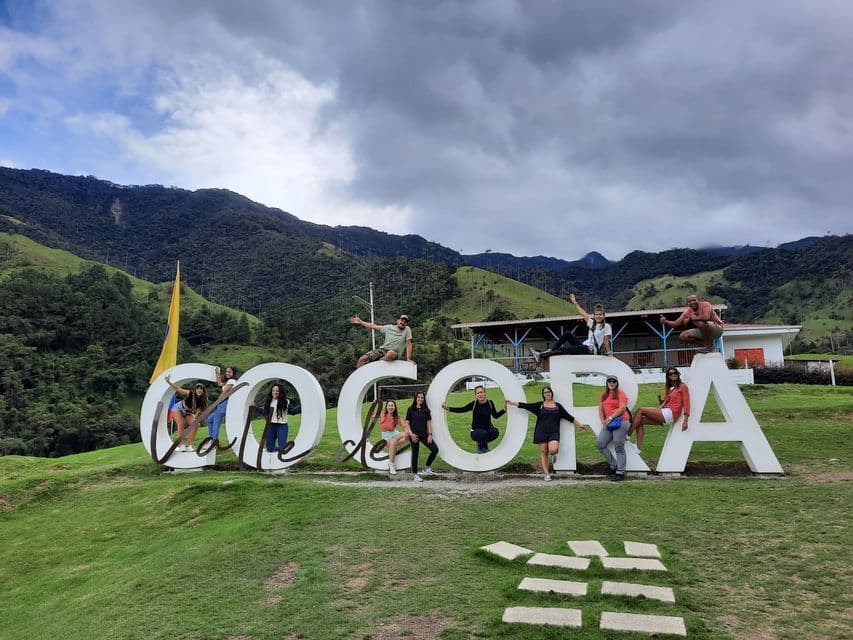 A WeRoad group trip posing for a photo on the large white 'Valle de Cocora' sign with a backdrop of green mountains.