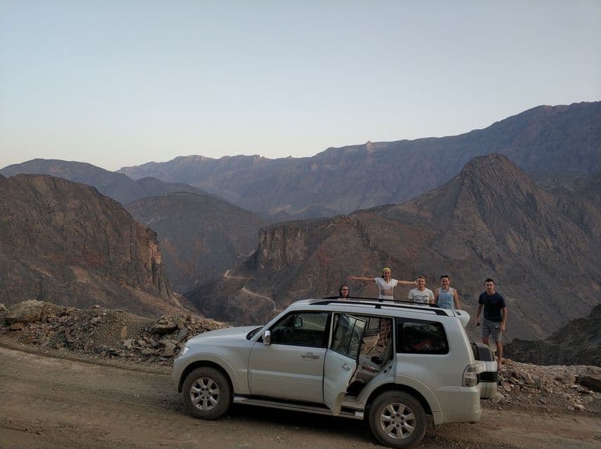Un groupe WeRoad en voyage pose devant un SUV blanc sur une piste de terre, surplombant une vaste chaîne de montagnes rocheuses.
